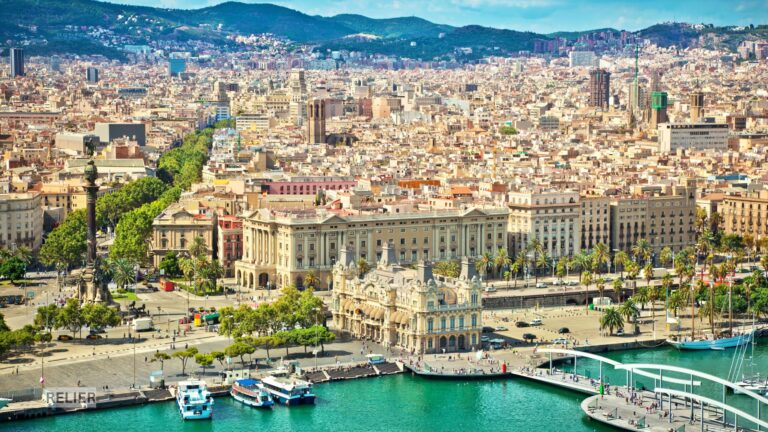 Aerial panoramic view of Barcelona showing the city’s grid layout, landmark architecture, and Mediterranean coastline under a clear sky.