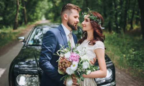 Newlyweds near the black wedding car stand on the road in the summer forest. A bride with a wreath of real flowers on her head.
